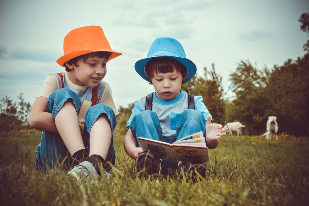 Twee kinderen met hoedjes die samen een boek lezen in het gras